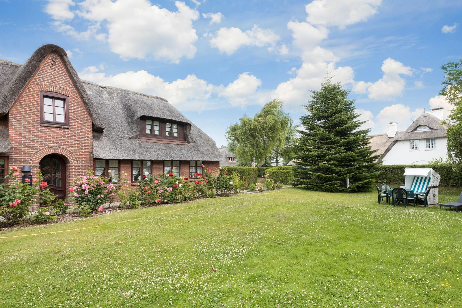 Haus mit Strohdach, Rasenfläche und Blumenbeete. Gartenmöbel und Nadelbaum sind sichtbar.