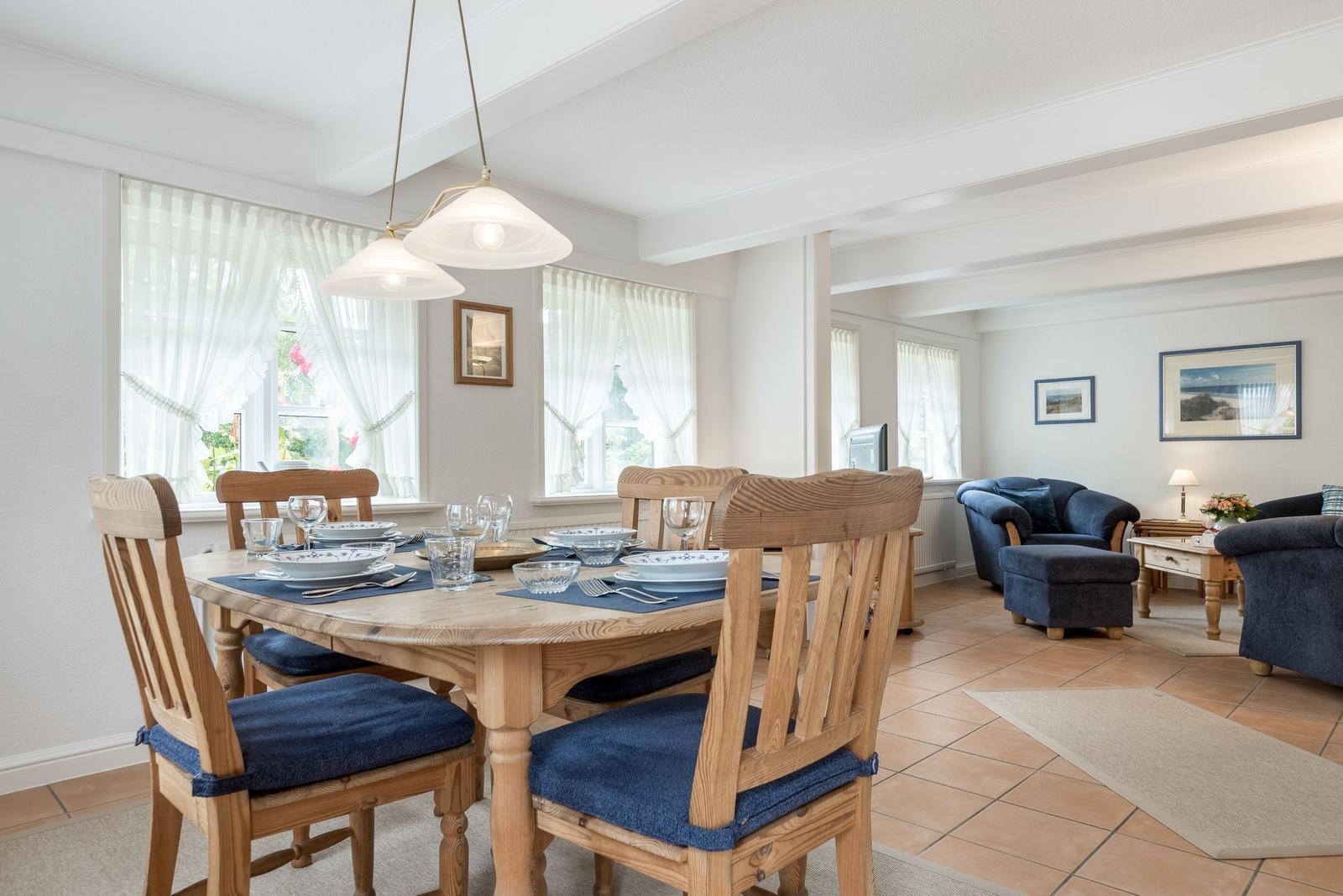 Esszimmer mit Holztisch und Stühlen, Wohnbereich mit blauen Sesseln und Fenstern mit weißen Vorhängen.