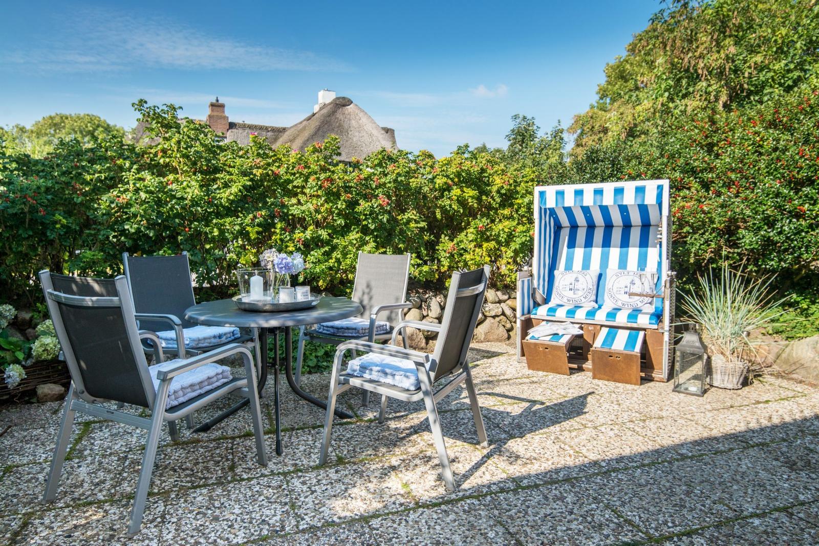 Terrasse mit Tisch, Stühlen und Strandkorb. Hintergrund: Grünflächen und Dachhäuser.