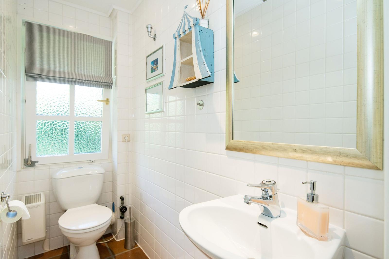 Badezimmer mit Toilette, Waschbecken und Spiegel. Fenster mit Rollladen und Wandregal.
