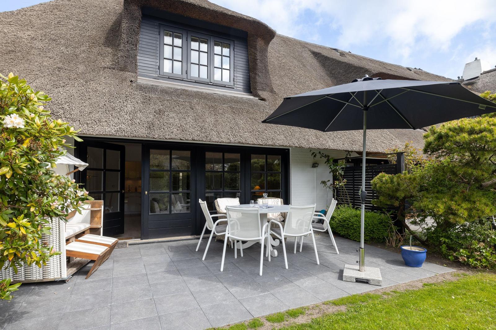 Terrasse mit Tisch, Stühlen und Sonnenschirm vor einem Haus mit Strohdach.