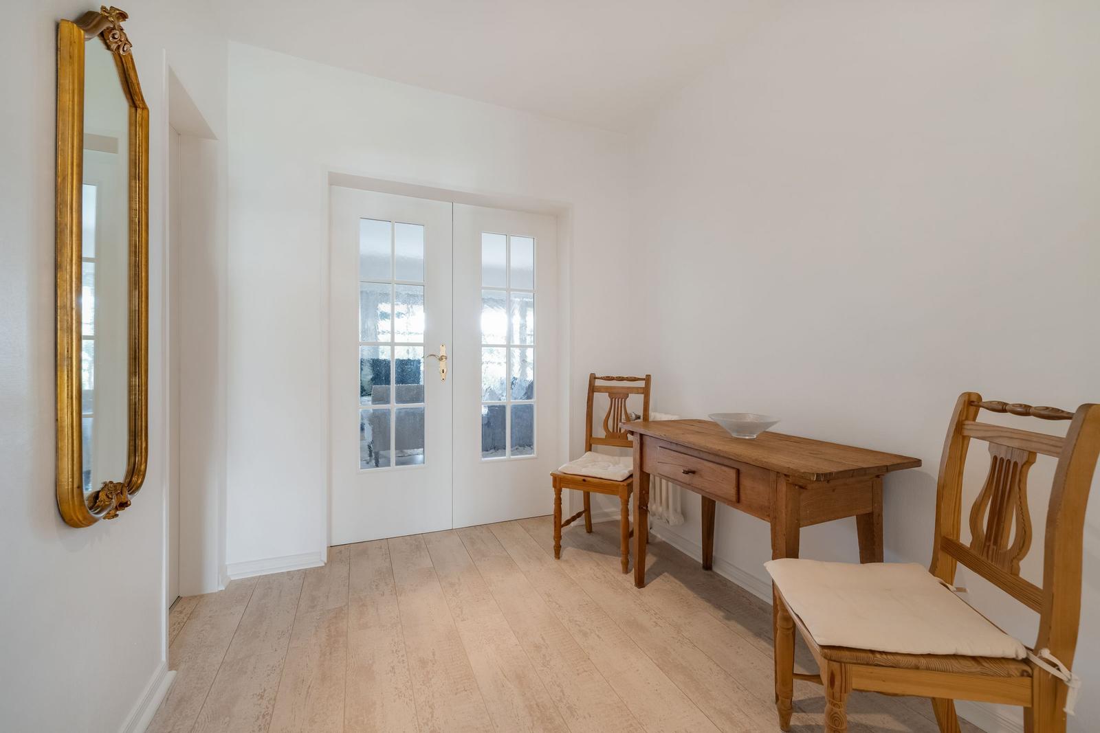 Raum mit Holztisch, zwei Stühlen und goldgerahmtem Spiegel an der Wand.