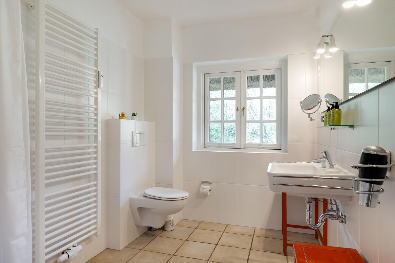 Badezimmer mit Toilette, Waschbecken und Fenster. Weiße Fliesen und Holzboden.