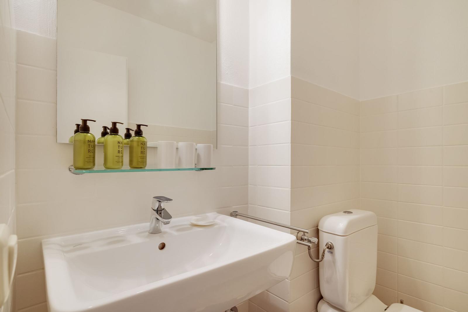 Badezimmer mit Waschbecken, Spiegel und Toilette. Auf dem Regal stehen grüne Flaschen.