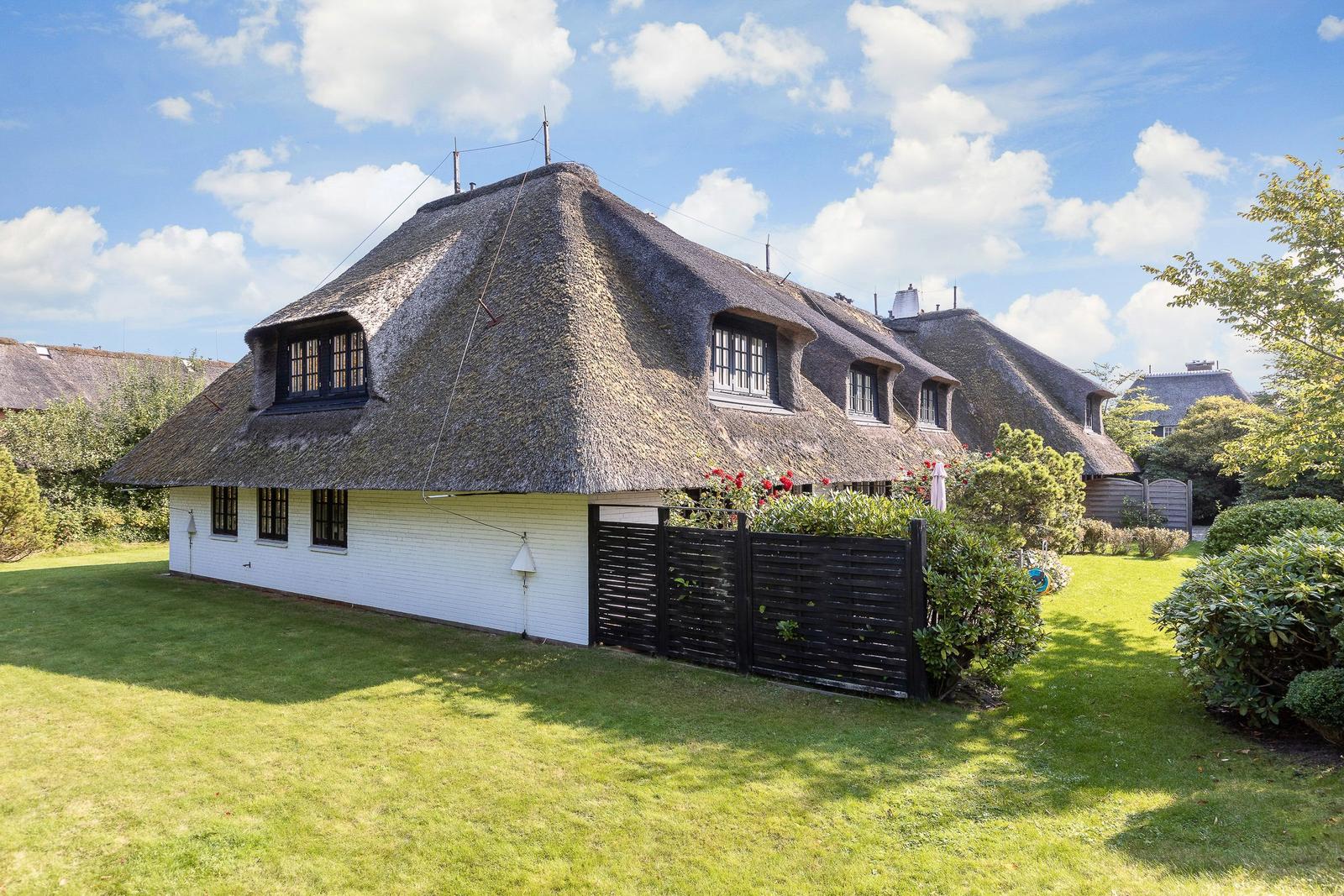 Haus mit strohgedecktem Dach und weißer Fassade, umgeben von Grünfläche und Hecken.