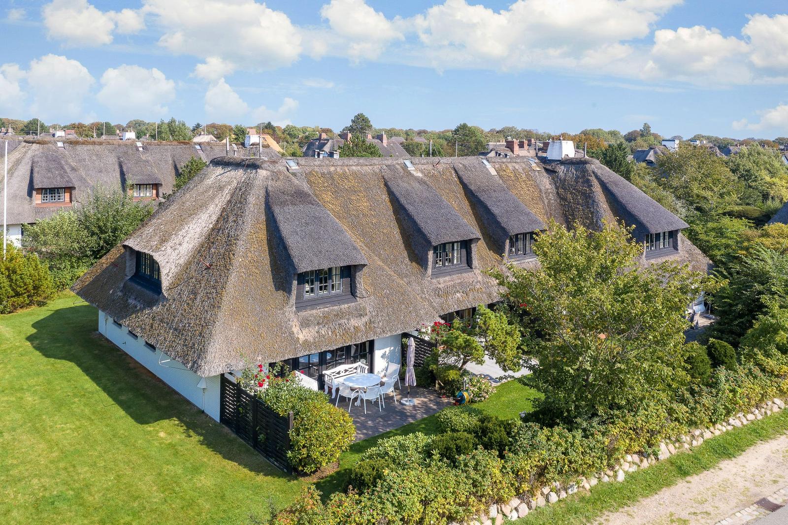 Haus mit Strohdach, Terrasse und Garten. Umgeben von Bäumen und Gras.
