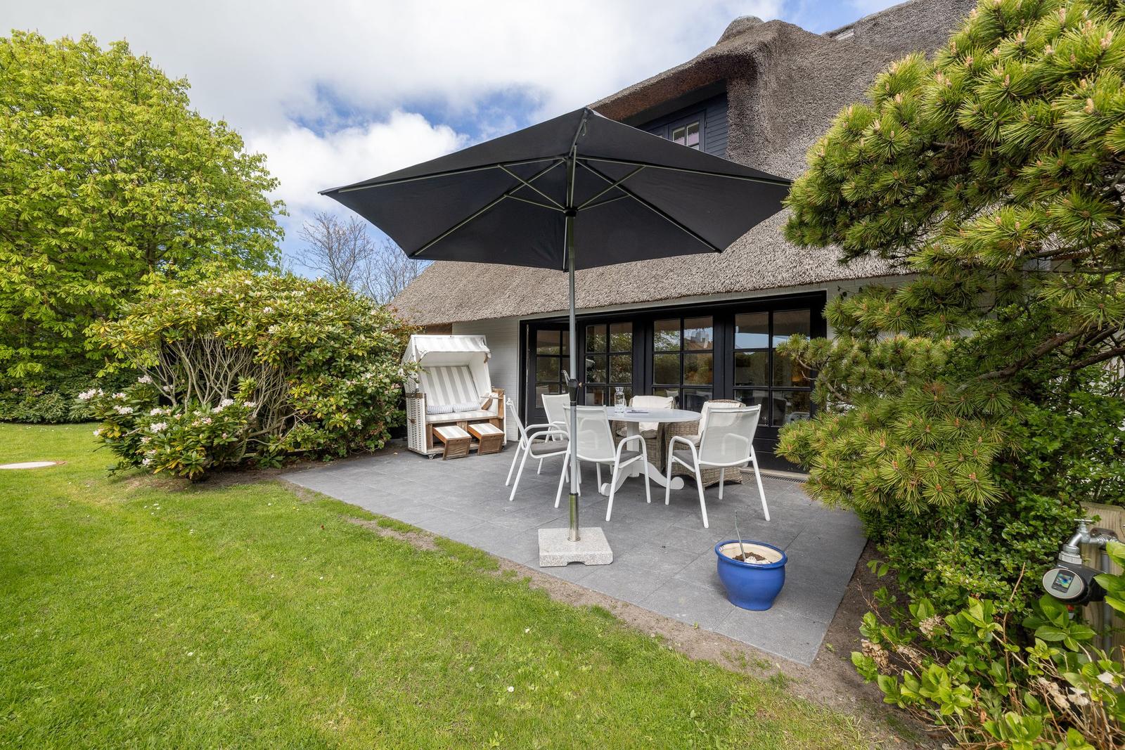 Terrasse mit Tisch, Stühlen und Sonnenschirm neben einem Haus mit Strohdach.