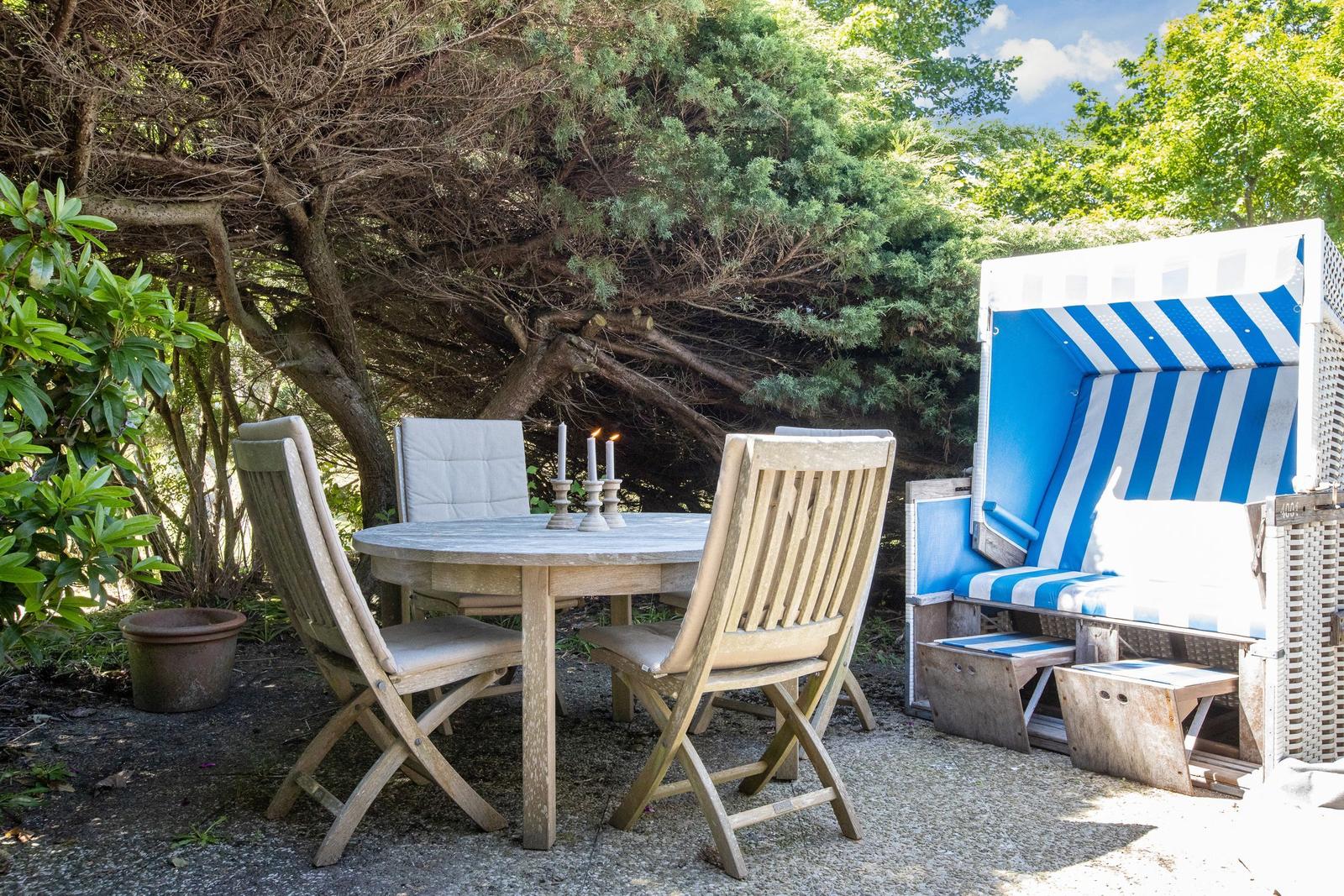 Holztisch mit Stühlen und Strandkorb im Garten unter Bäumen.
