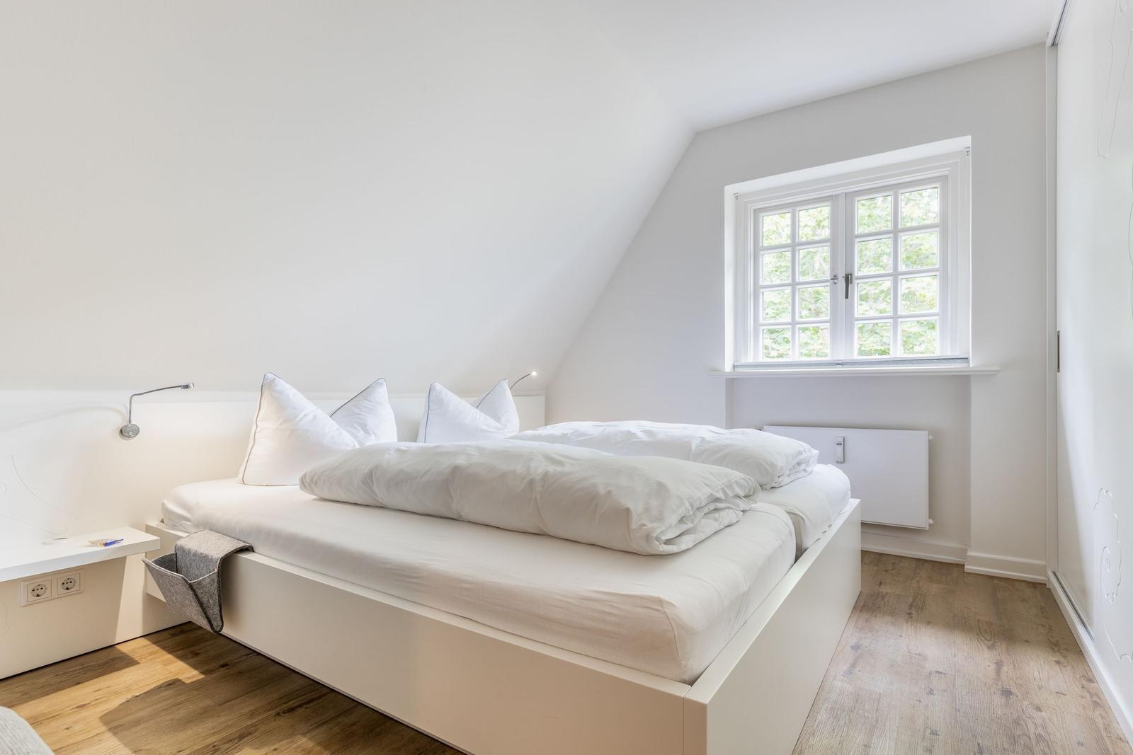 Schlafzimmer mit weißem Bett, Fenster und Holzboden.