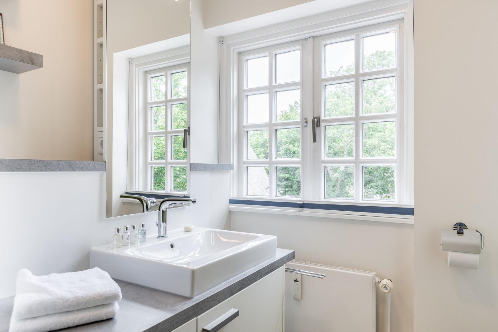 Badezimmer mit Waschbecken, Spiegel und Fenster mit Blick auf Bäume.