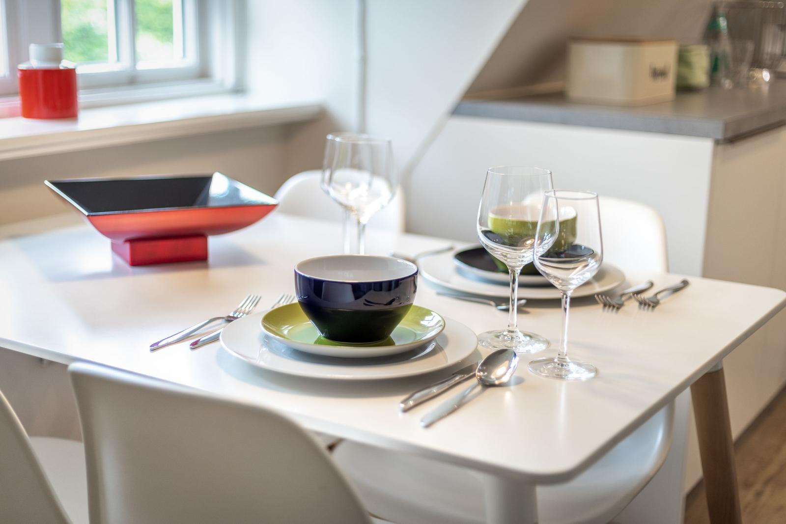 Tisch mit Geschirr und Gläsern, Fenster und Küchenbereich im Hintergrund.