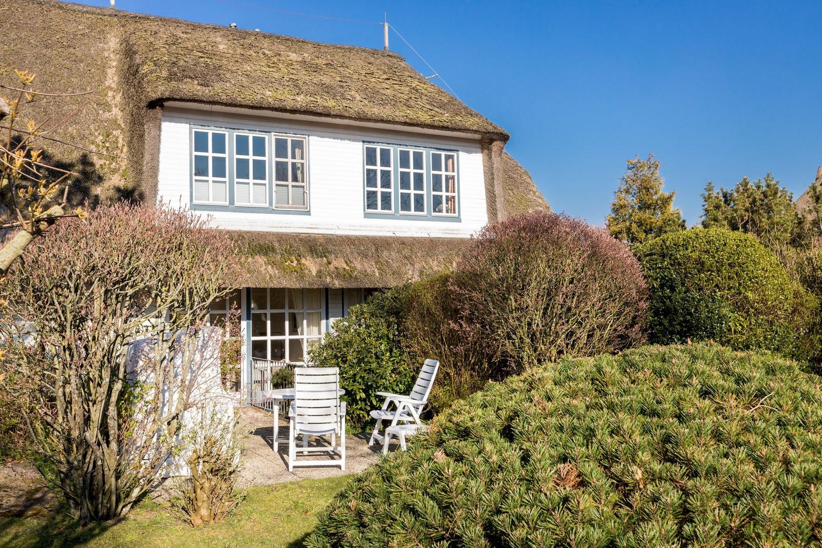 House with thatched roof, white facade, and garden with white chairs.
