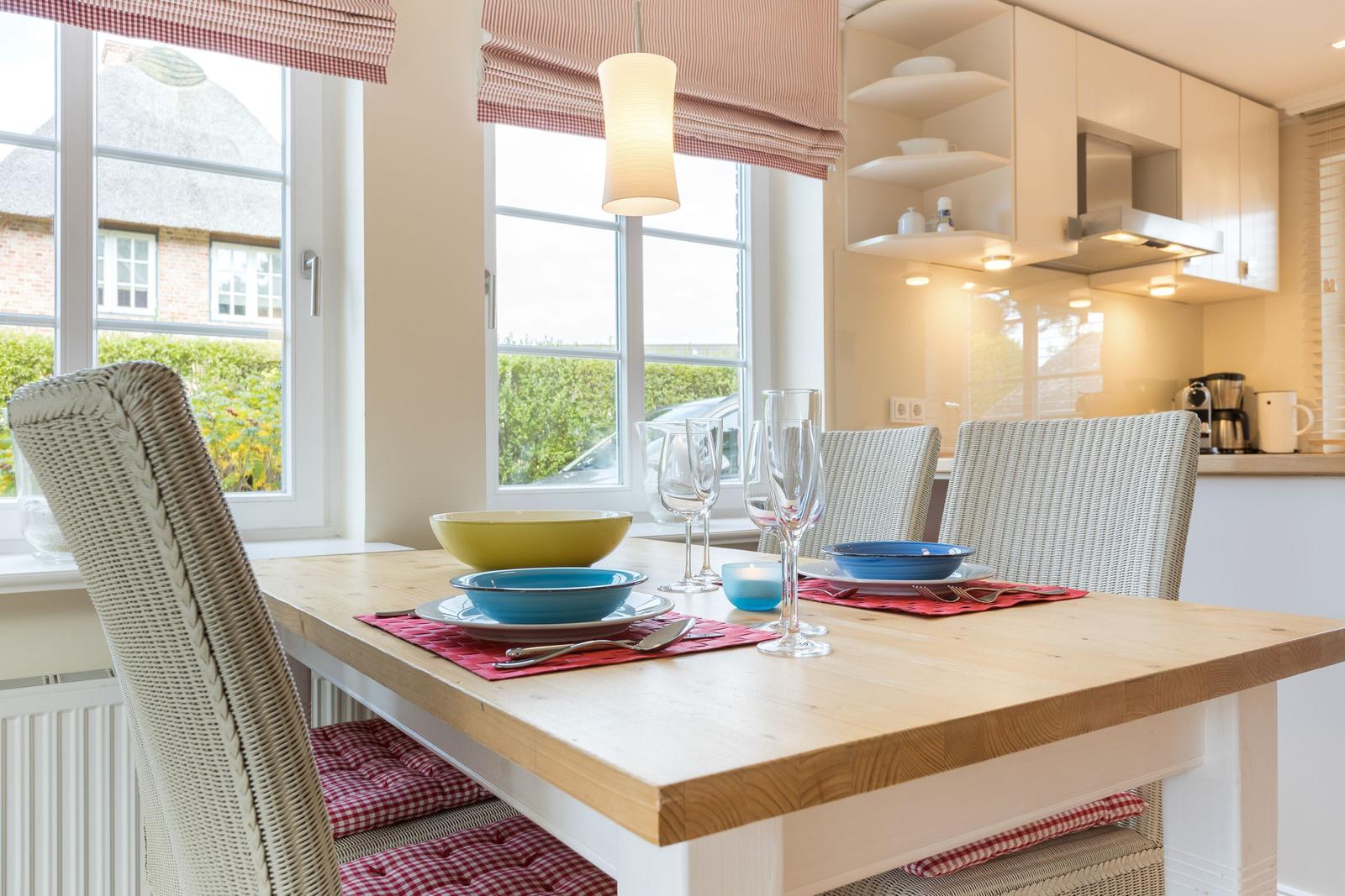 Dining area with wooden table, chairs, and windows. Kitchen in background.