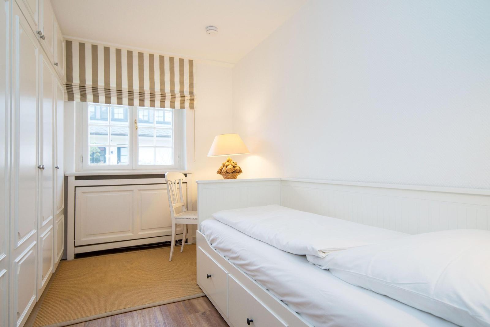 Schlafzimmer mit weißem Bett, Kleiderschrank und Fenster mit Markise.