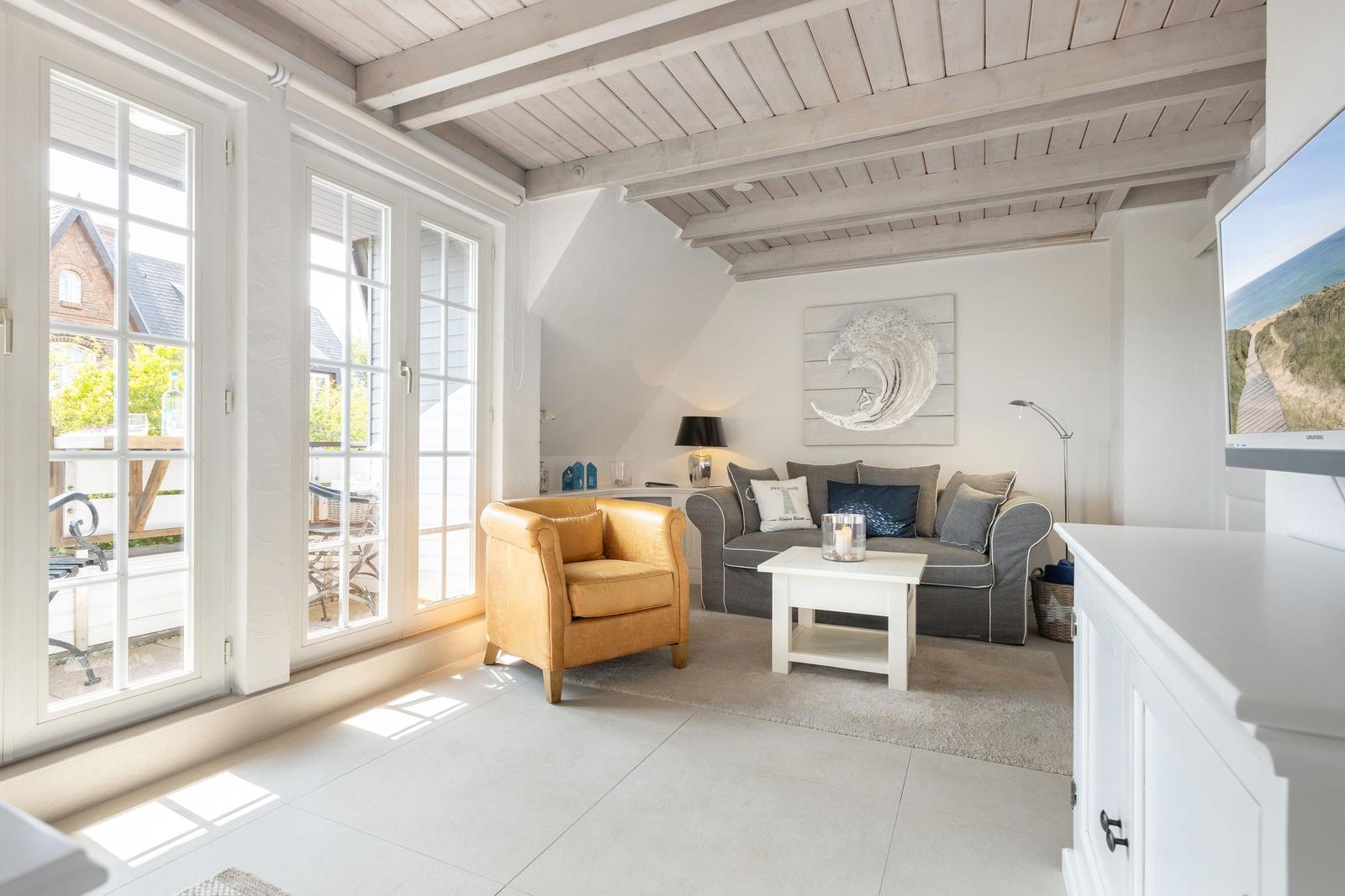 Wohnzimmer mit Sofa, Sessel und großen Fenstern. Decke mit Holzbalken und Wandbild.
