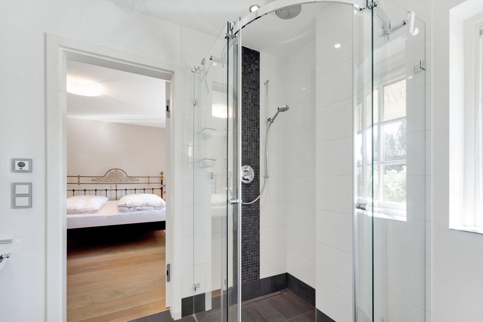 Shower with glass enclosure and view into bedroom with bed.