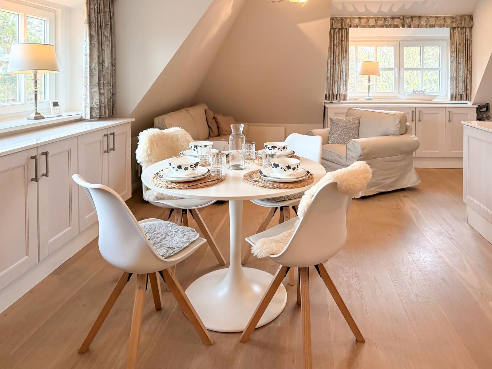 Dining room with round table, white chairs, and windows. Wooden floor and light furnishings.
