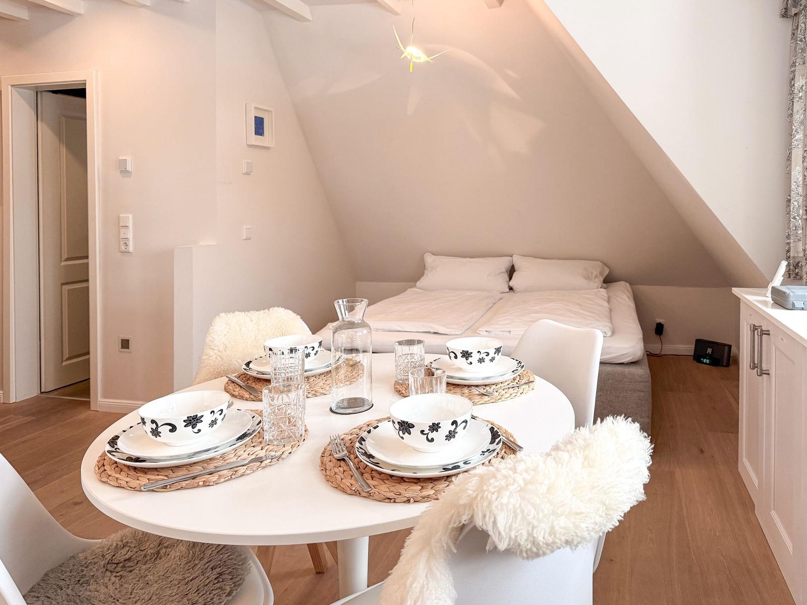 Round table with dishes, white chairs, and sleeping area under gable roof.