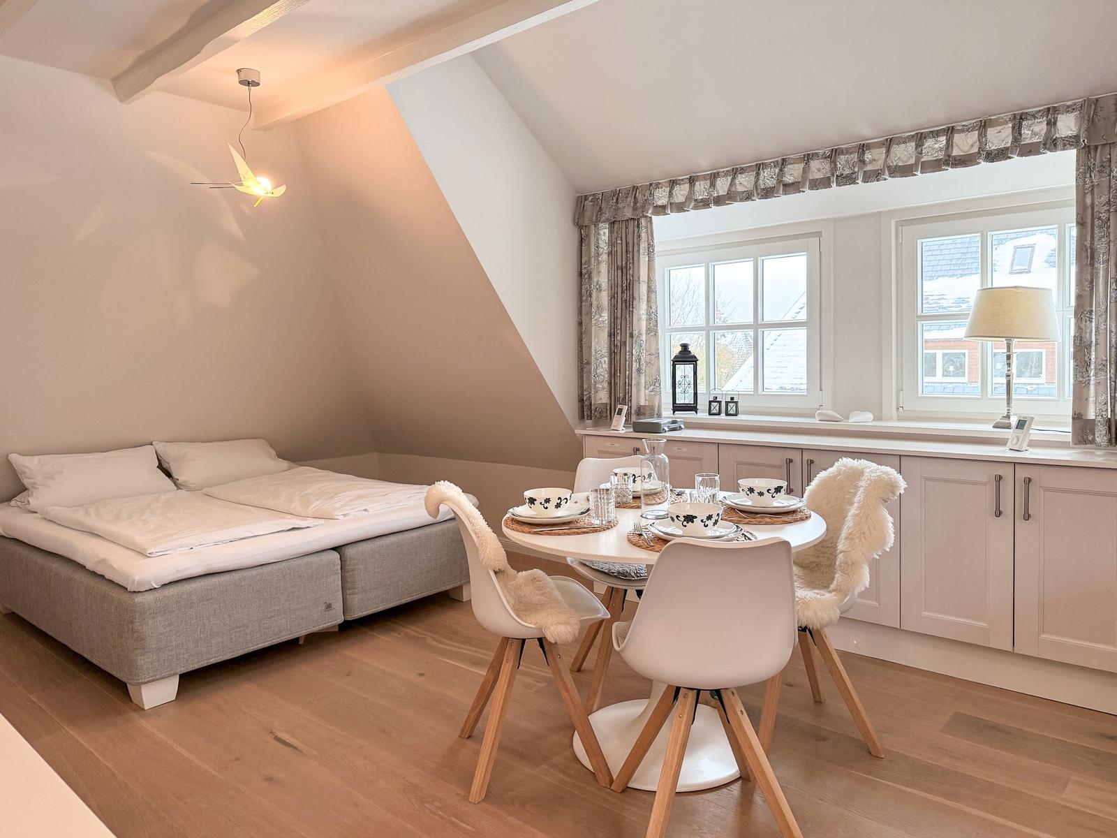 Bedroom with double bed and dining table with four chairs. Wooden floor and large windows.