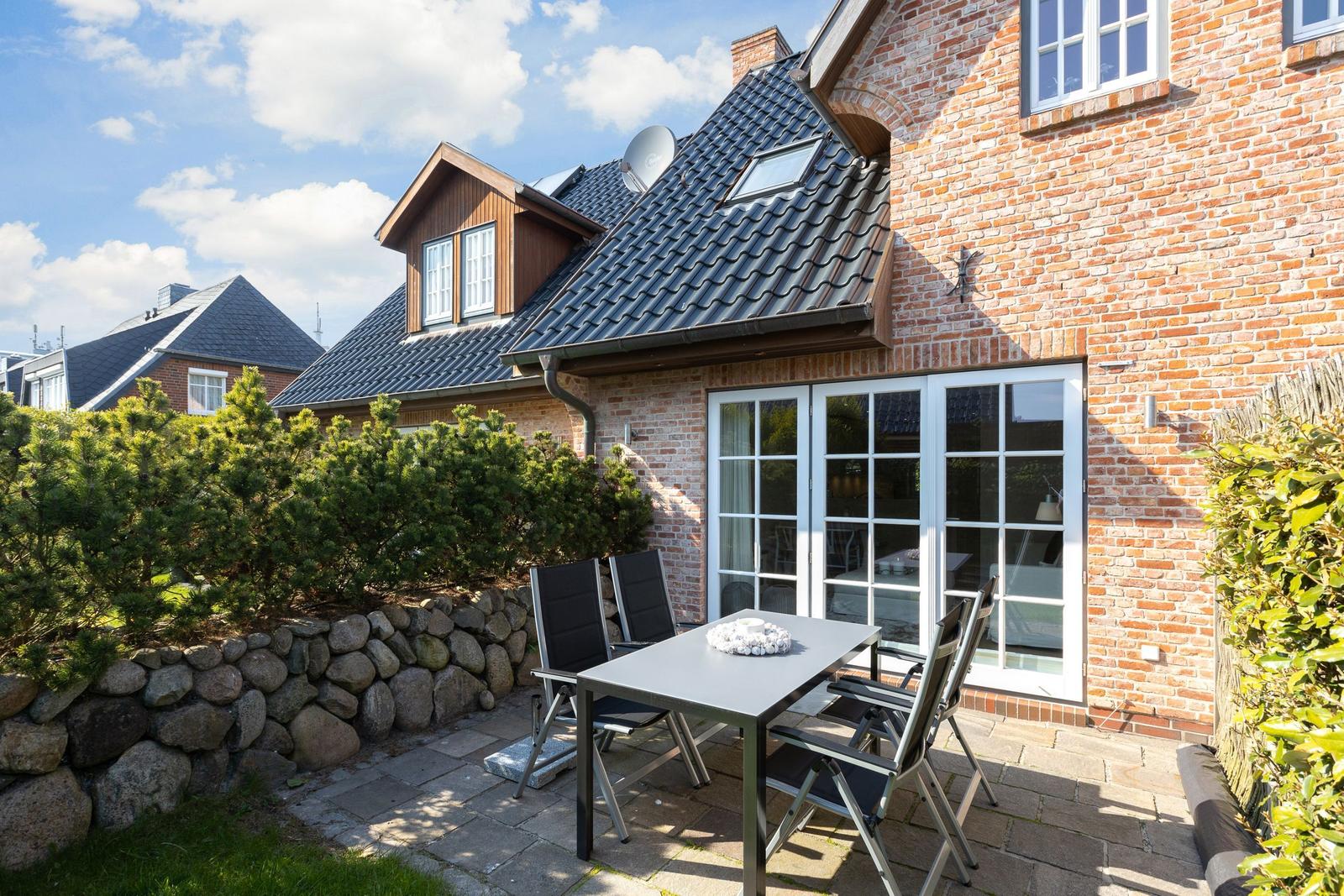 Terrace with table and chairs in front of a house with brick wall and glass doors.