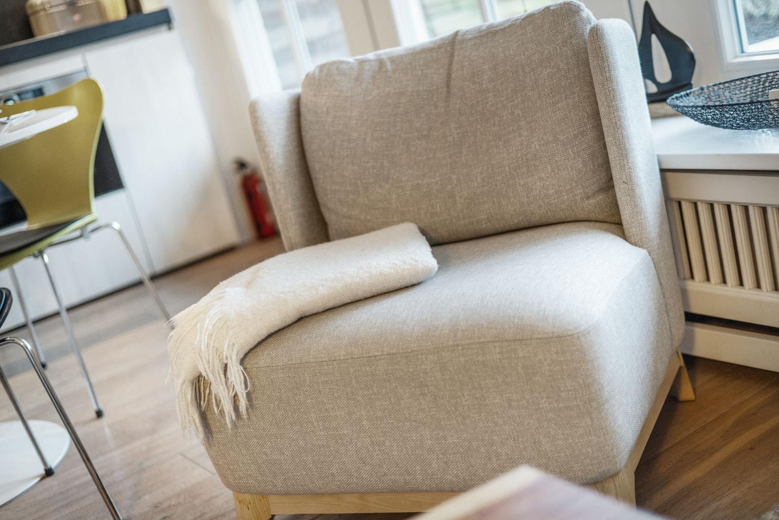 Ein bequemer Sessel mit einer Decke auf dem Sitz. Im Hintergrund ist eine Küche mit Stuhl zu sehen.