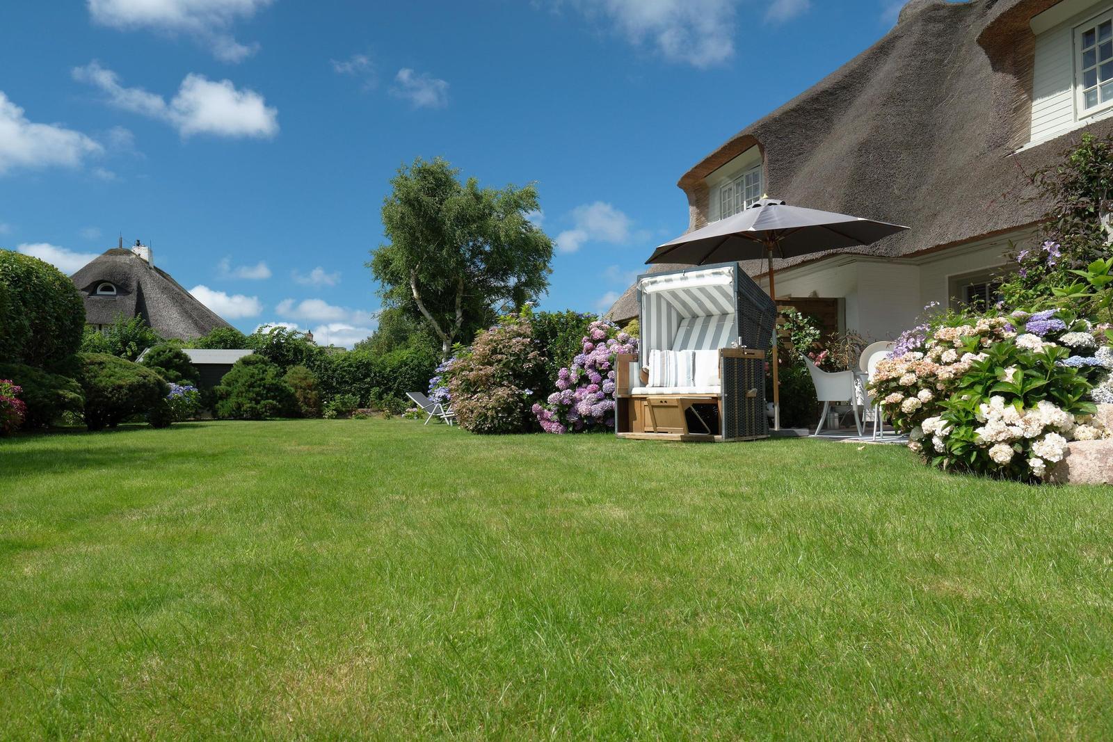 Grüner Garten mit Strandkorb und Sonnenschirm vor einem Haus mit Strohdach.