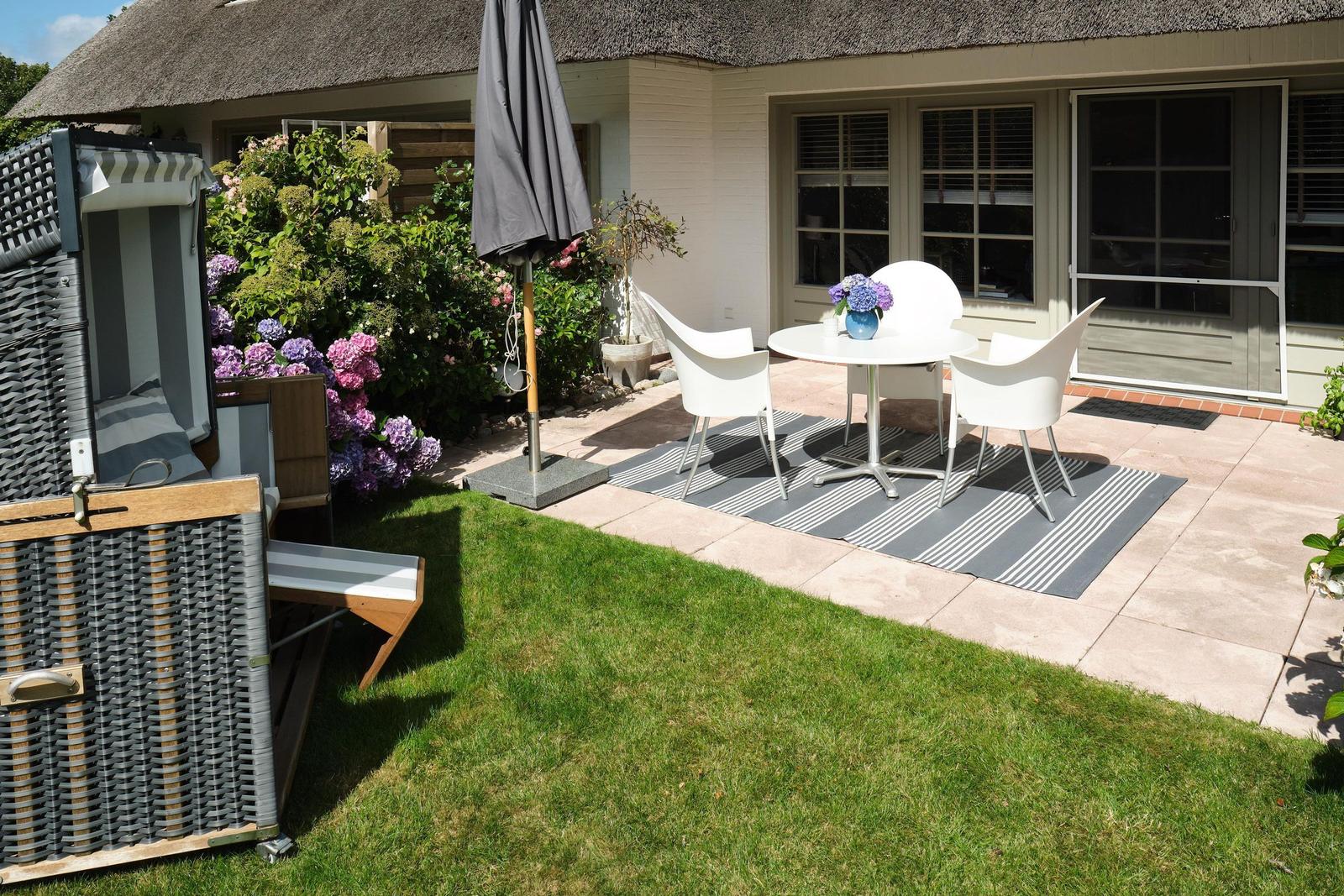 Terrasse mit weißen Stühlen, Tisch und Sonnenschirm neben einem Gartenhaus.