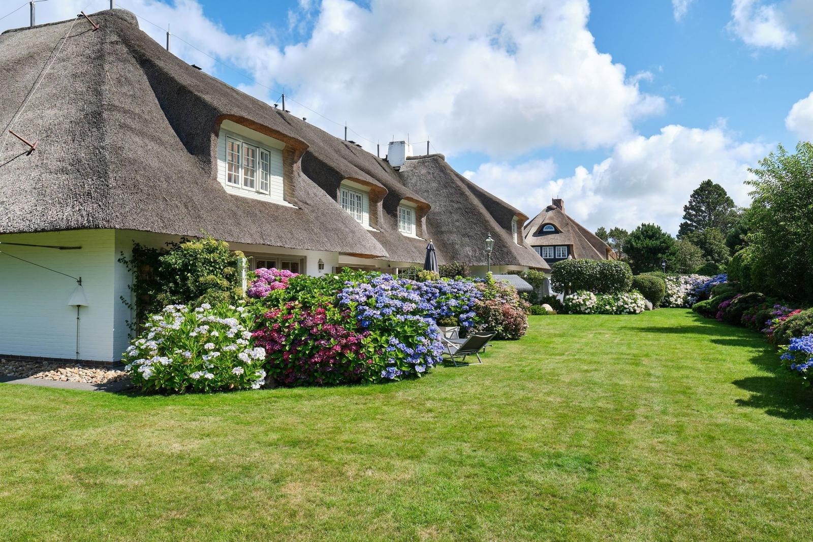 Haus mit Strohdach, Blumenbeete und Rasenfläche unter blauem Himmel.