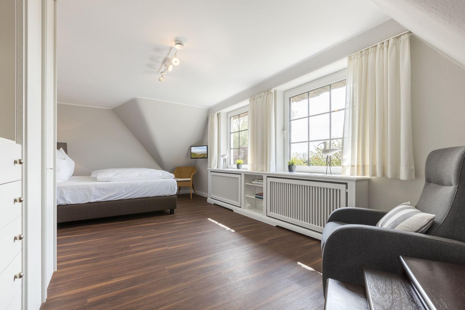Schlafzimmer mit Bett, Sessel und Fenstern mit Vorhängen.