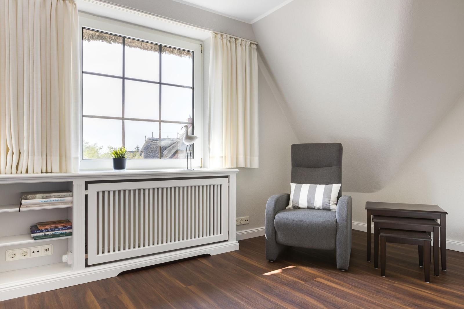 Ein Raum mit grauem Sessel, Fenster und Holzboden. Neben dem Fenster steht ein weißer Schrank mit Bücherregal.