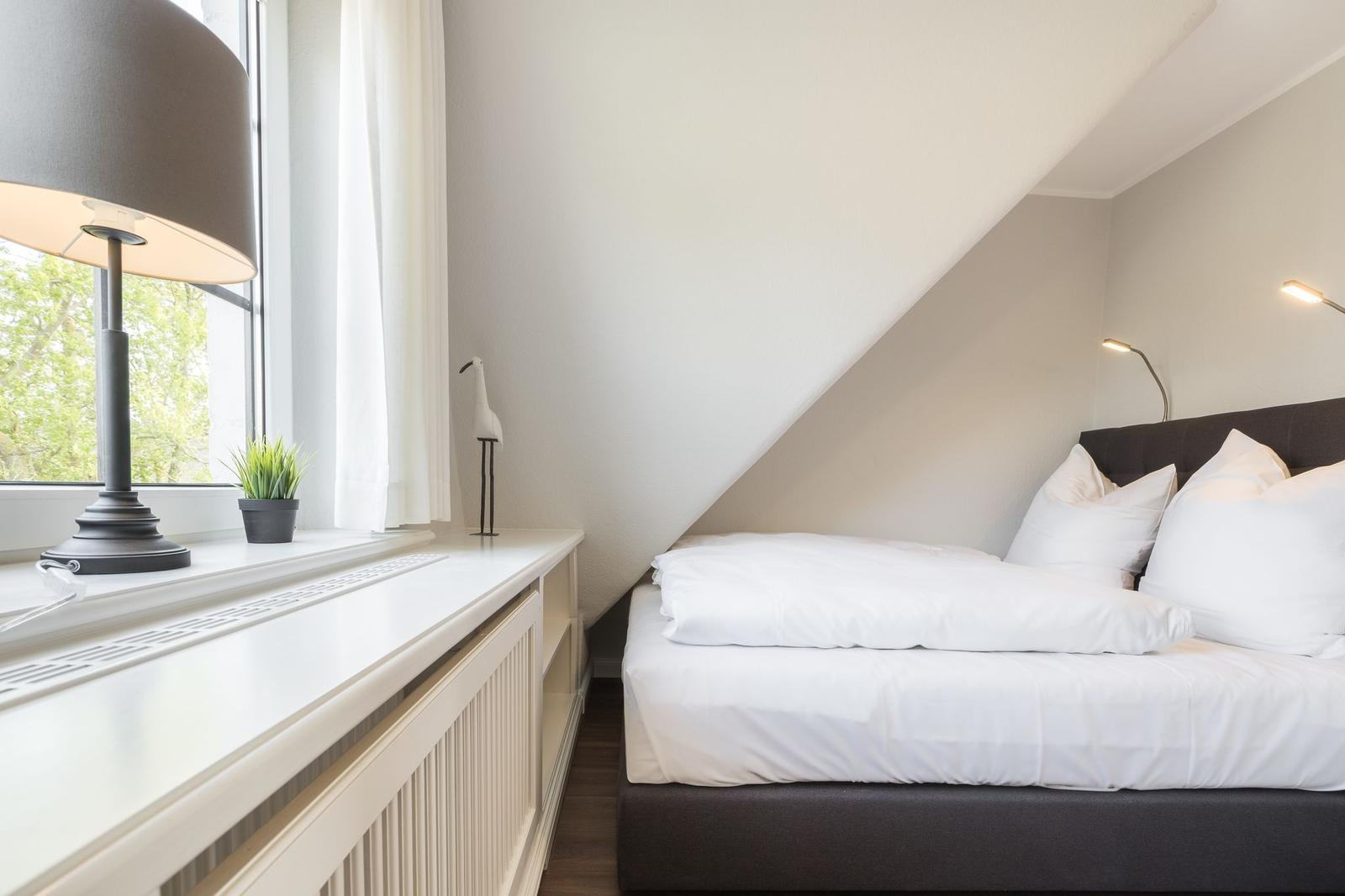 Schlafzimmer mit Dachschräge, Bett und Fensterbank mit Lampe und Pflanze.