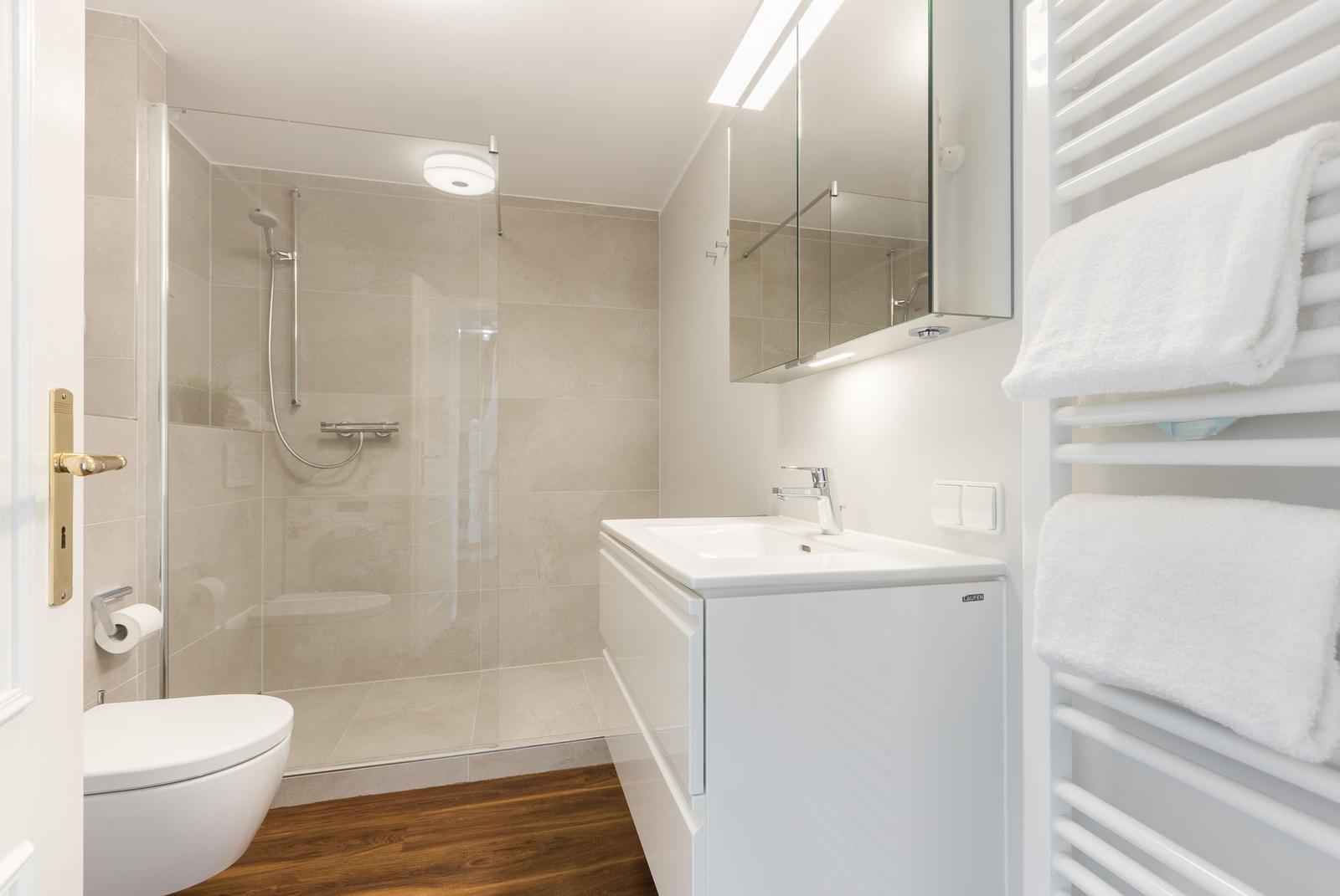 Badezimmer mit Dusche, Waschbecken und Heizung. Holzboden und weiße Fliesen.