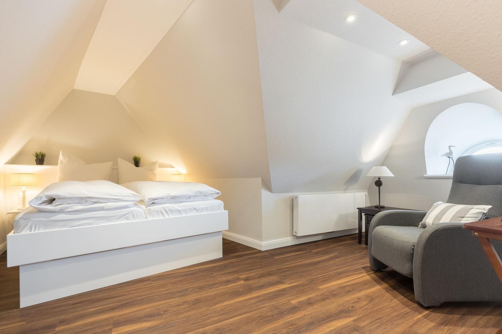 Schlafzimmer mit Dachschrägen, weißem Bett und grauem Sessel.