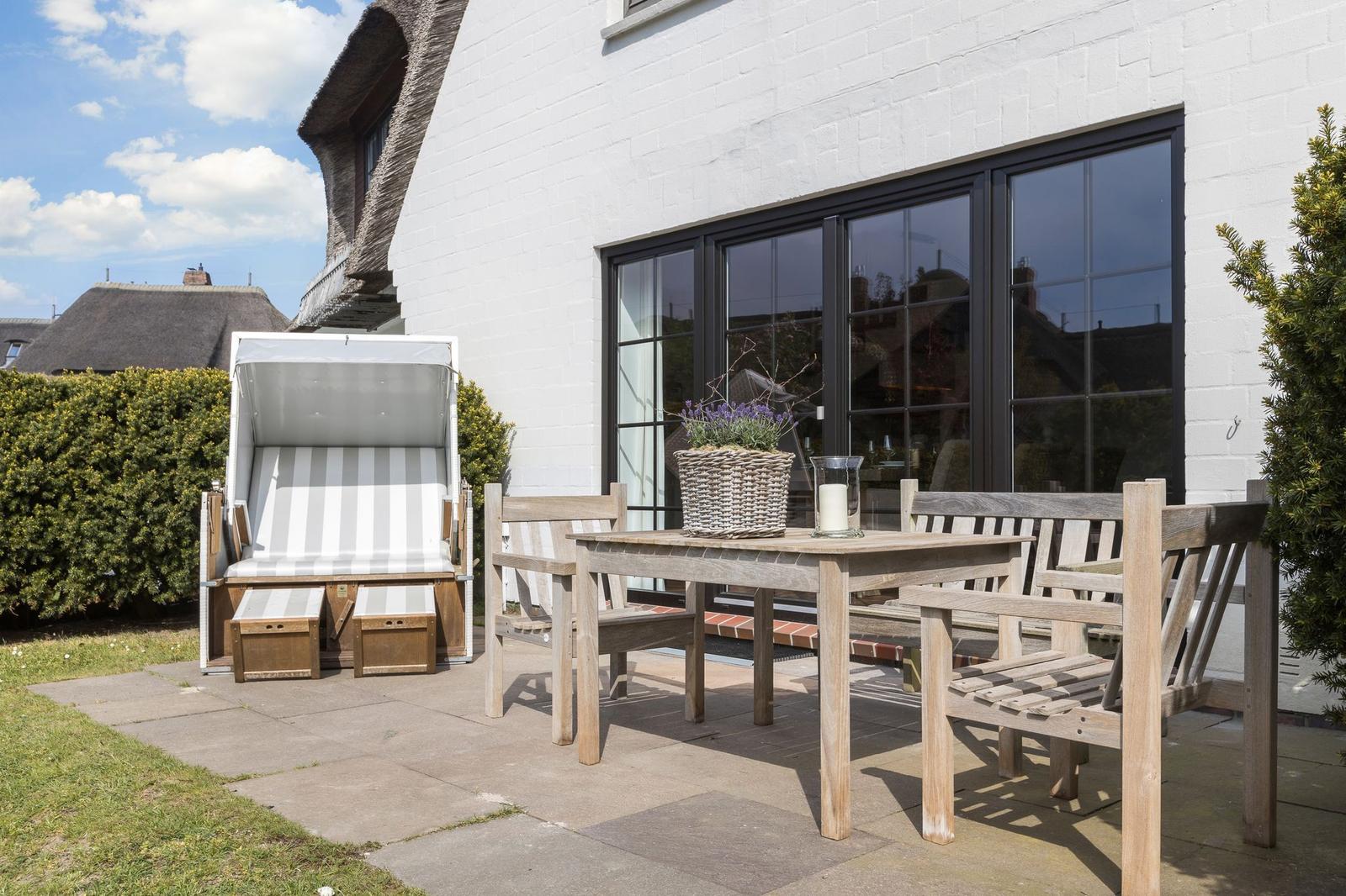 Terrasse mit Holztisch, Bänken und Strandkorb. Hintergrund: weißer Hauswand mit Fenstern.