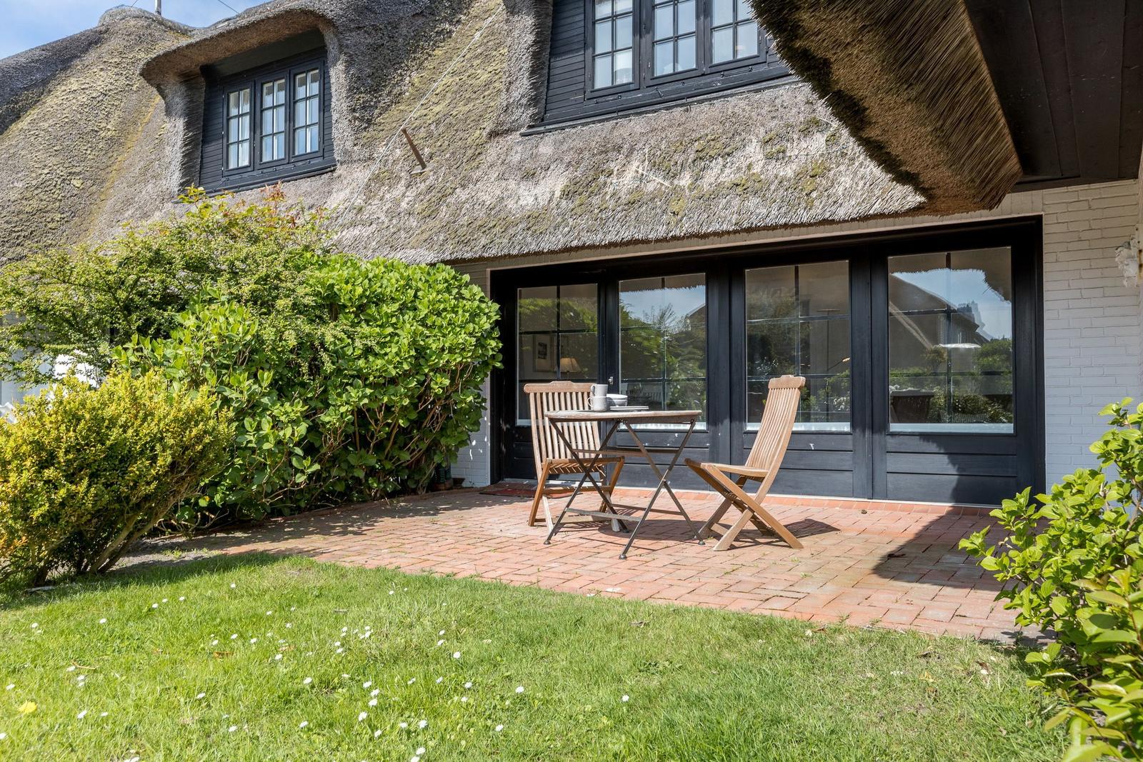 Terrasse mit Tisch und zwei Stühlen vor einem Haus mit Strohdach und großen Fenstern.