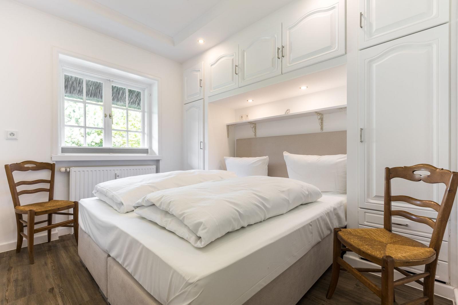 Schlafzimmer mit Doppelbett, weißen Schränken und Holzstühlen. Fenster mit Blick auf Grün.