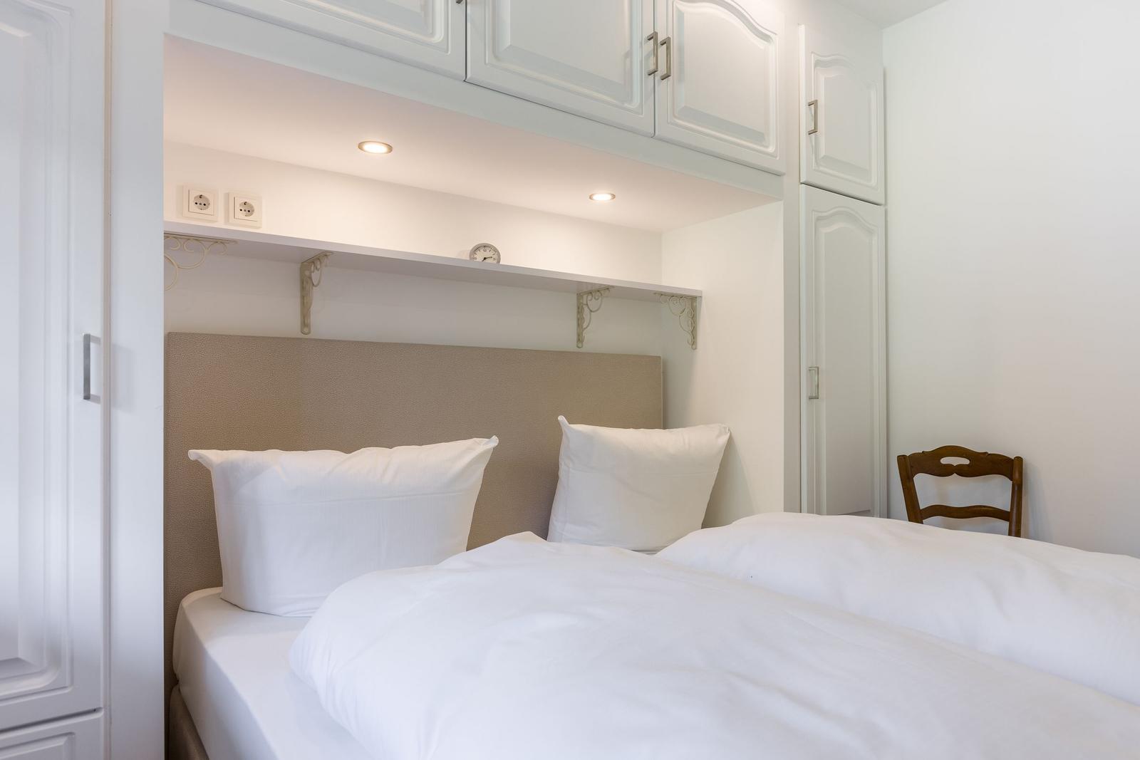 Schlafzimmer mit Bett, weißen Kissen und Holzstuhl. Oberhalb des Bettes befinden sich Schränke und eine Ablage.