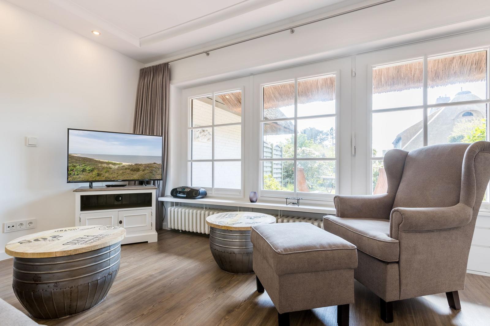 Ein Wohnzimmer mit Fernseher, Sessel und Fenstern zum Blick ins Freie.