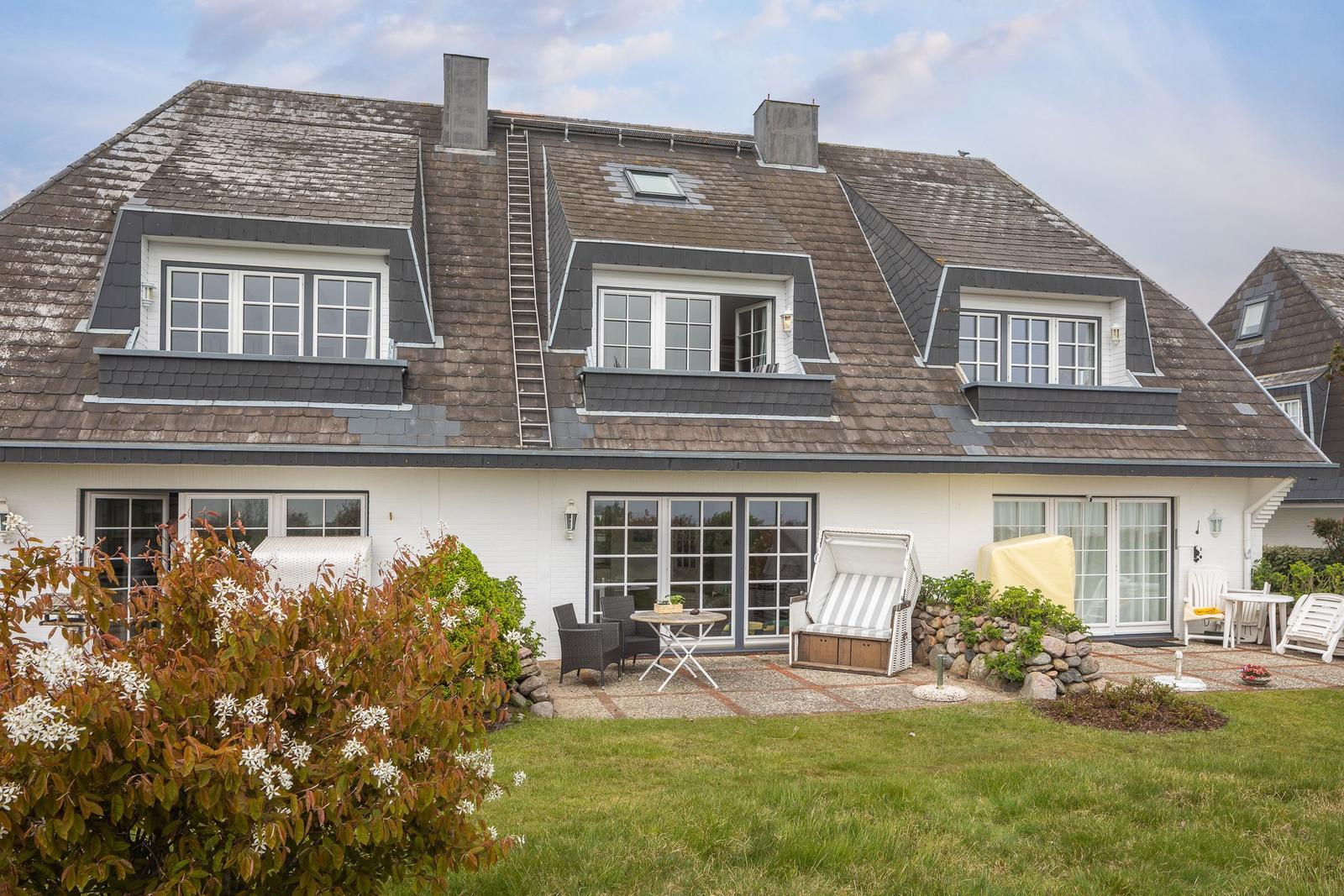 Haus mit Dachgauben, Terrasse und Garten. Weiße Fassade, Holzterrasse mit Möbeln.