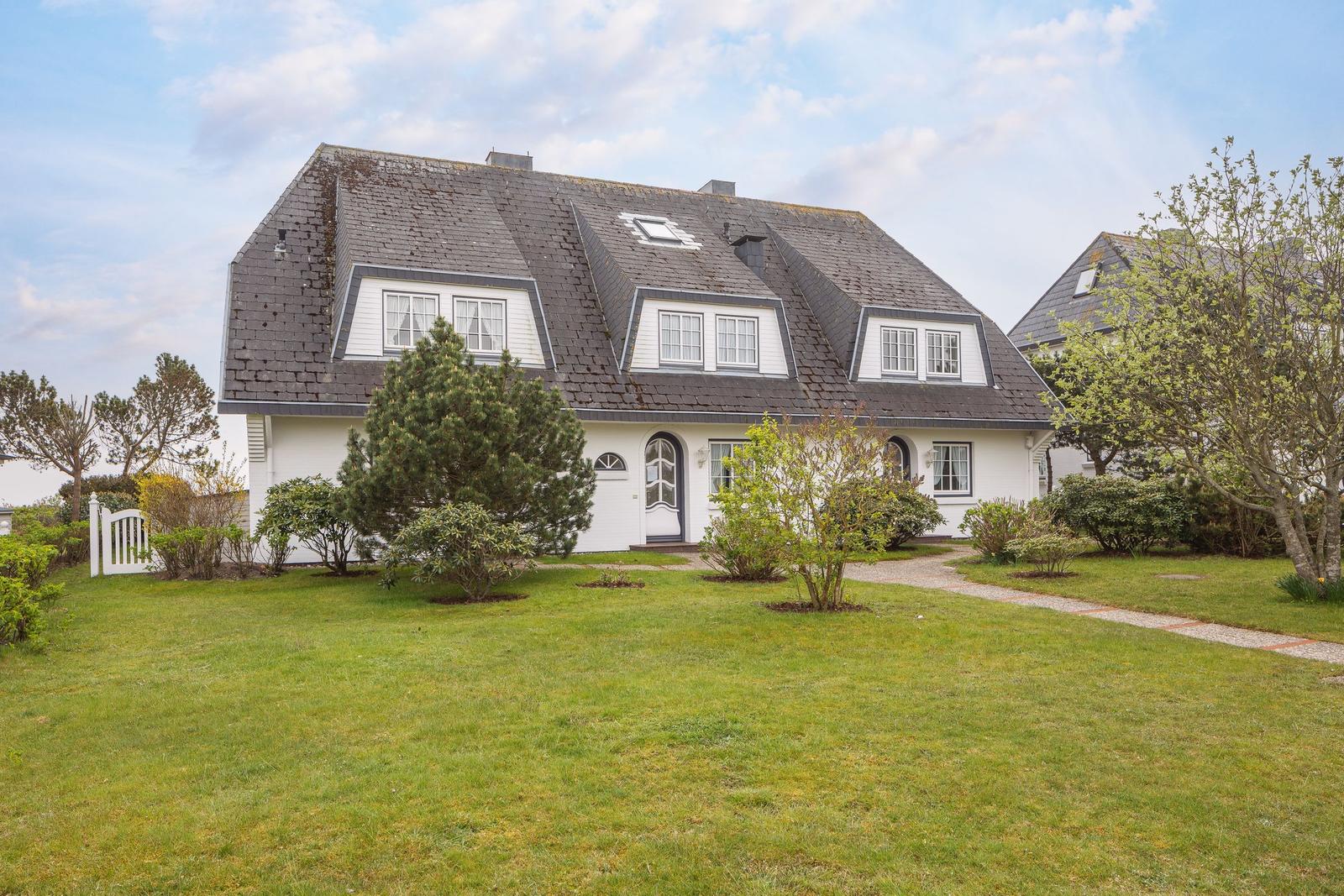 Wohnhaus mit grauem Dach und weißer Fassade, umgeben von Grünfläche und Bäumen.