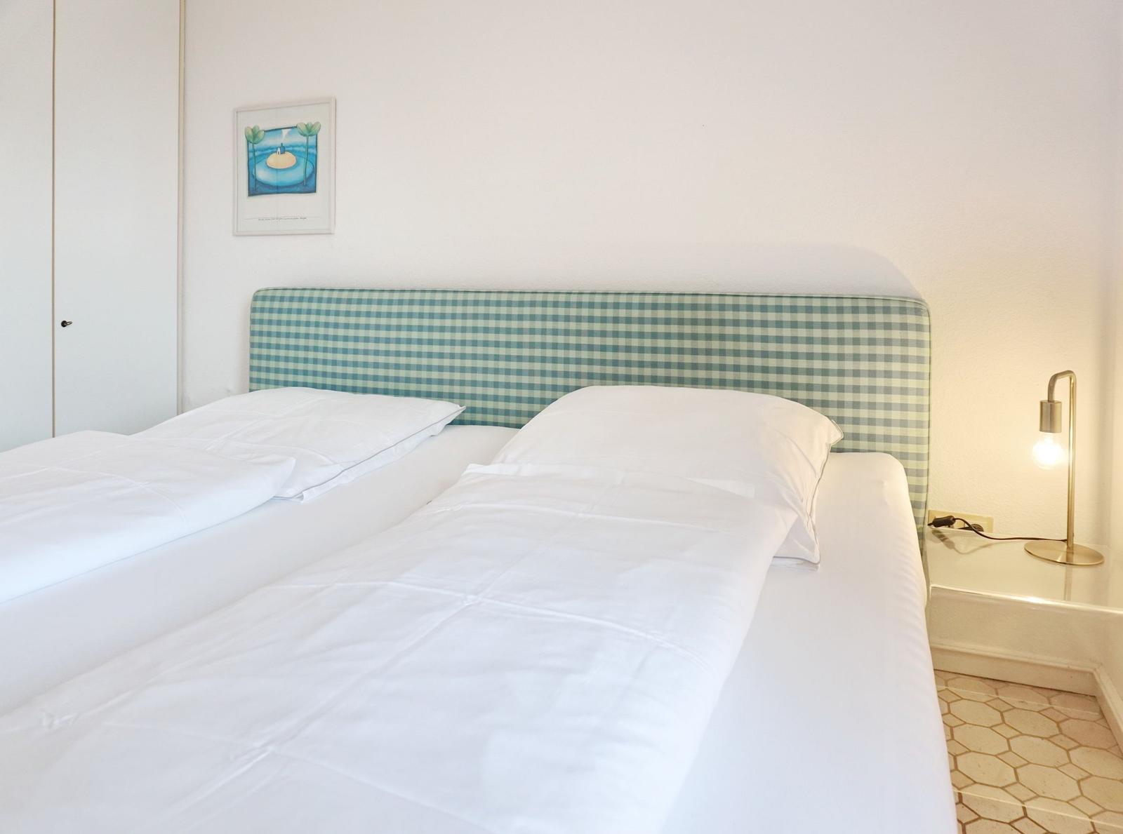 Double bed with white bedding and checkered headboard. Nightstand with lamp and wall art.