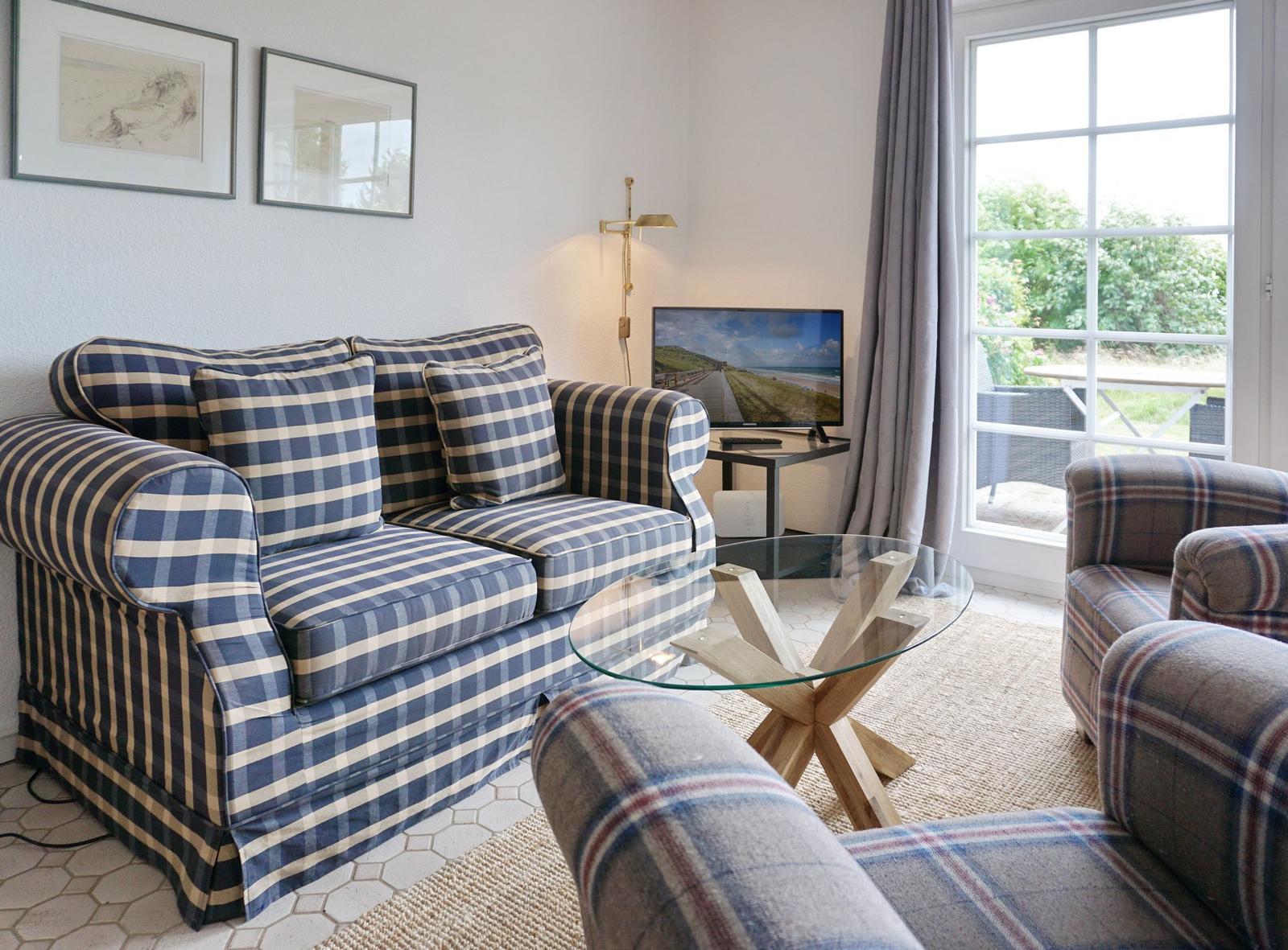 Living room with sofa, armchairs, glass table, and TV. View to balcony with garden.