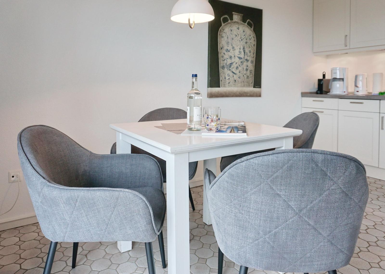 Dining room with white table and gray chairs, kitchen area in background.