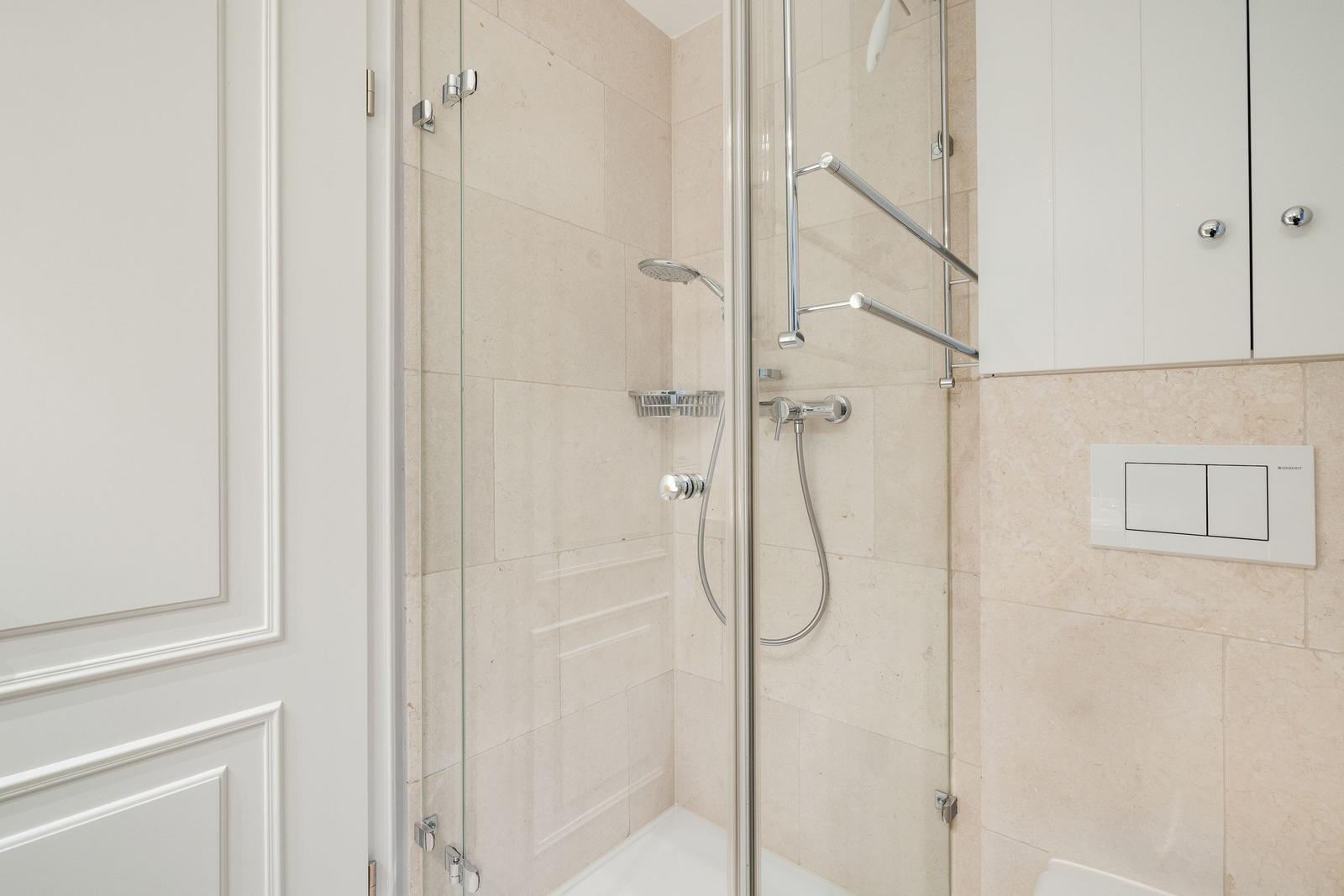 Shower with glass enclosure, showerhead, and handheld shower. White tiles and cabinet.