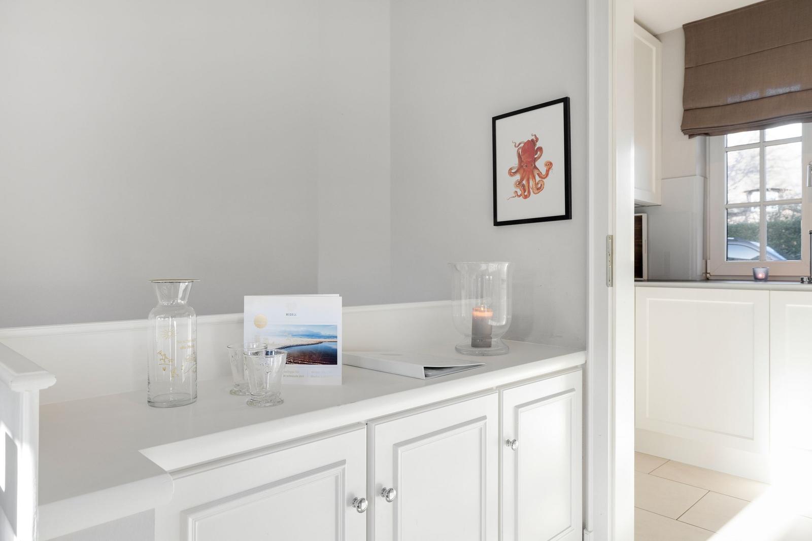 White cabinet with glass vases and picture on the wall. Kitchen in the background.