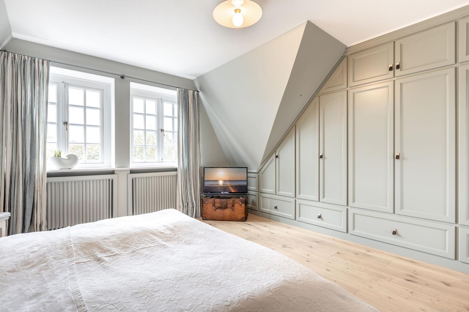 Schlafzimmer mit Dachschräge, großem Kleiderschrank und Fenstern mit Vorhängen.