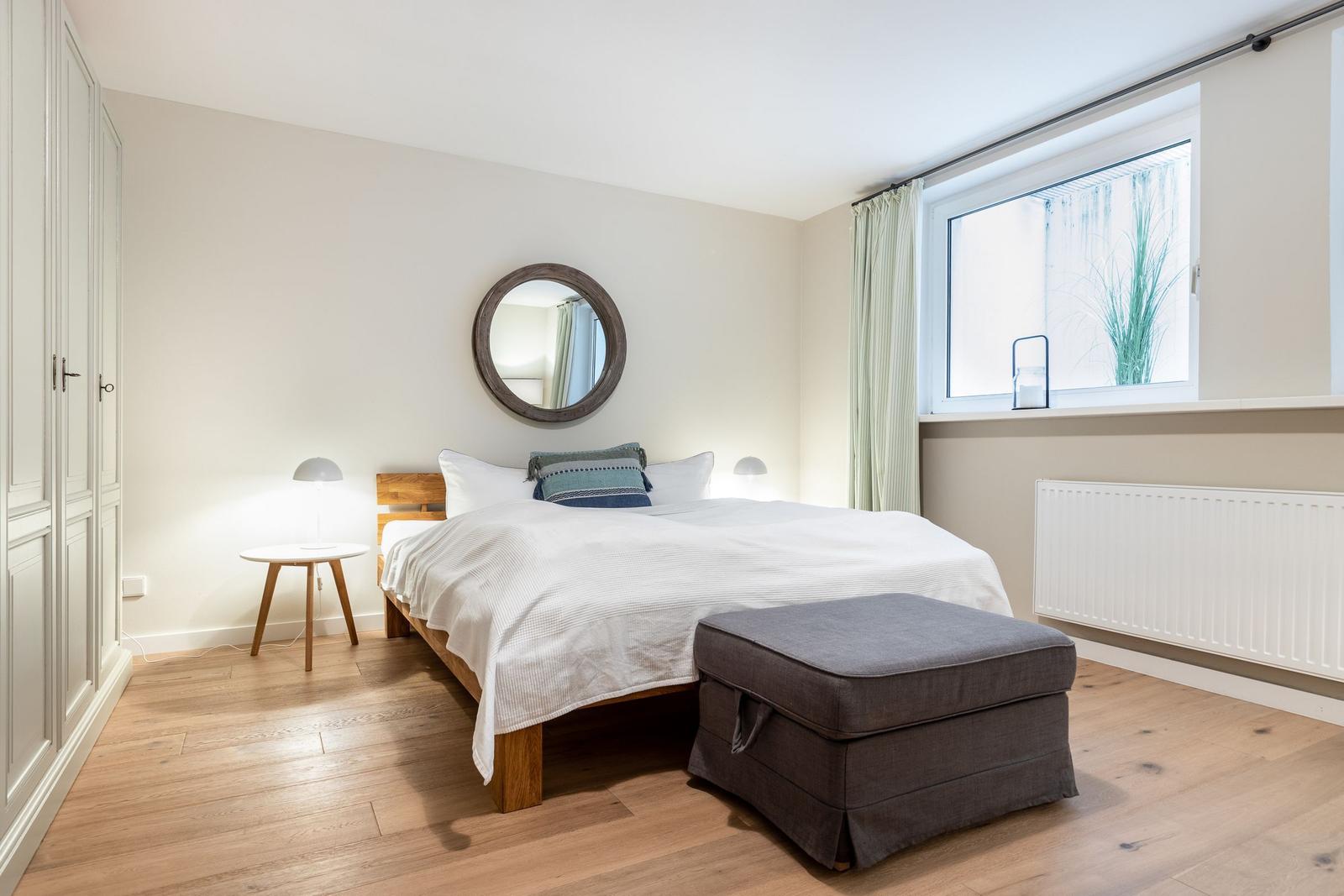 Schlafzimmer mit Bett, Nachttisch, Spiegel und Fenster.