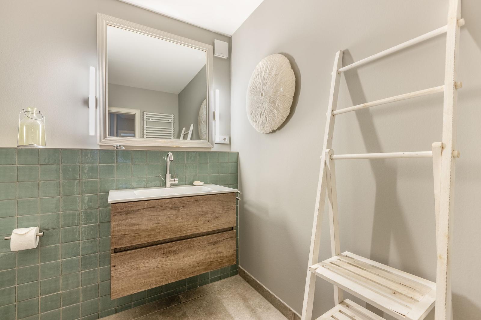 Badezimmer mit Waschbecken, Spiegel und Holzunterschrank. An der Wand hängt ein weißer Trockner.