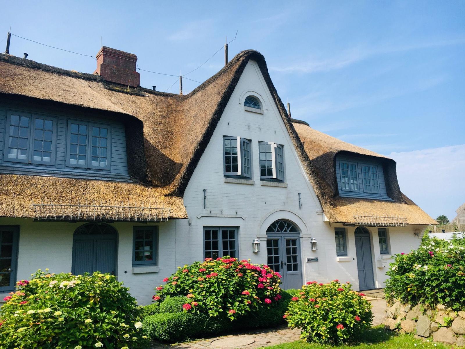 Haus mit Strohdach, weißen Wänden und blauen Fenstern, umgeben von Blumenbeeten.