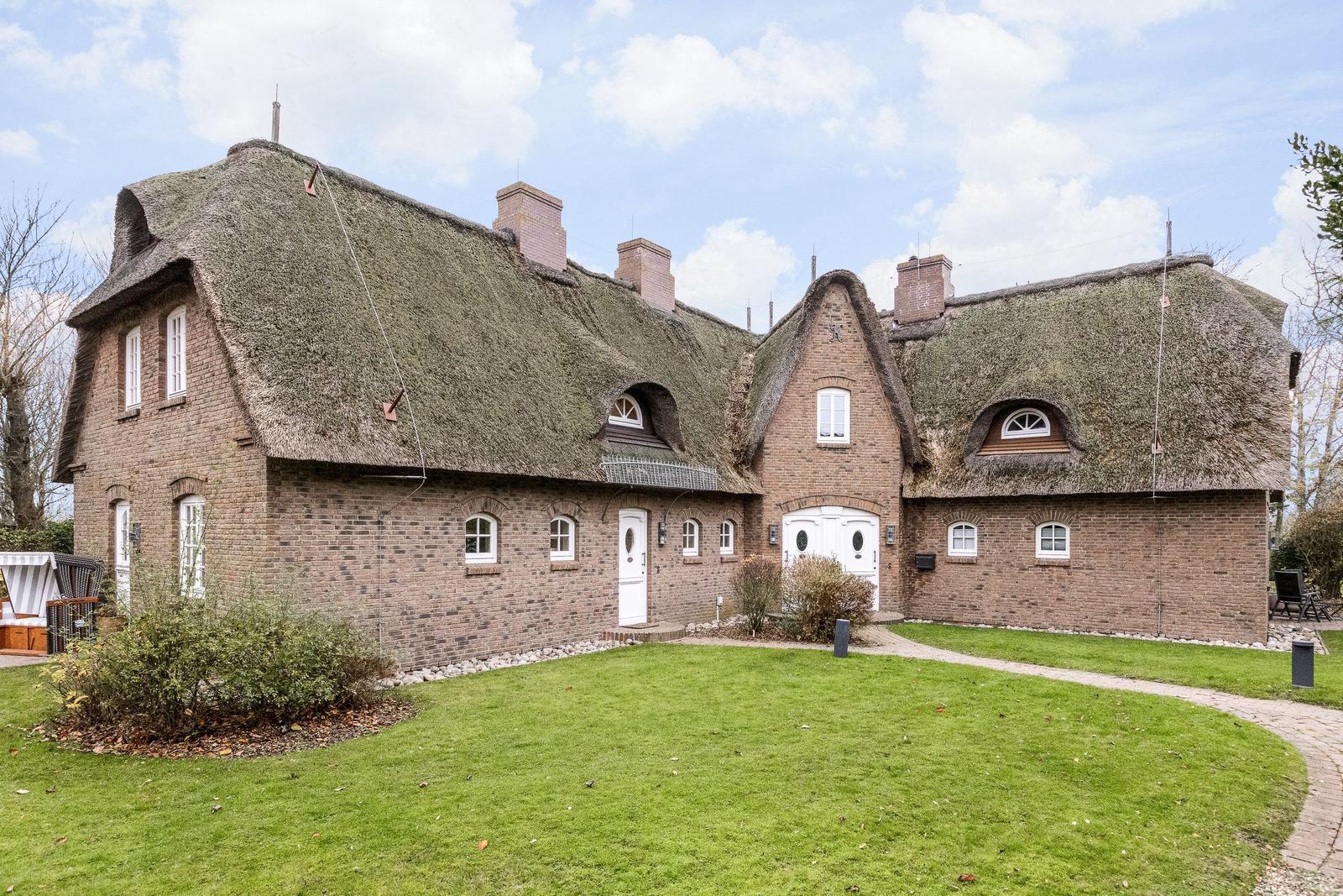 House with thatched roof and brown brick walls. Lawns and white doors.
